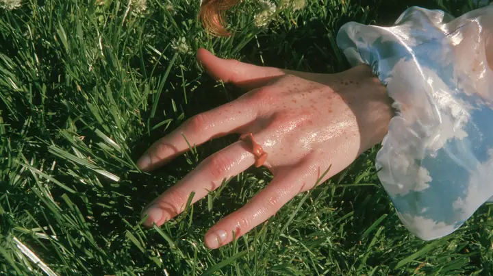 Y2K high fashion editorial, a 2001 couture magazine story: a macro of a pale freckled hand with a small clay-orange enamel ring brushing the tips of short bright-green grass, a sleeve of sky-print chiffon, sunlight…