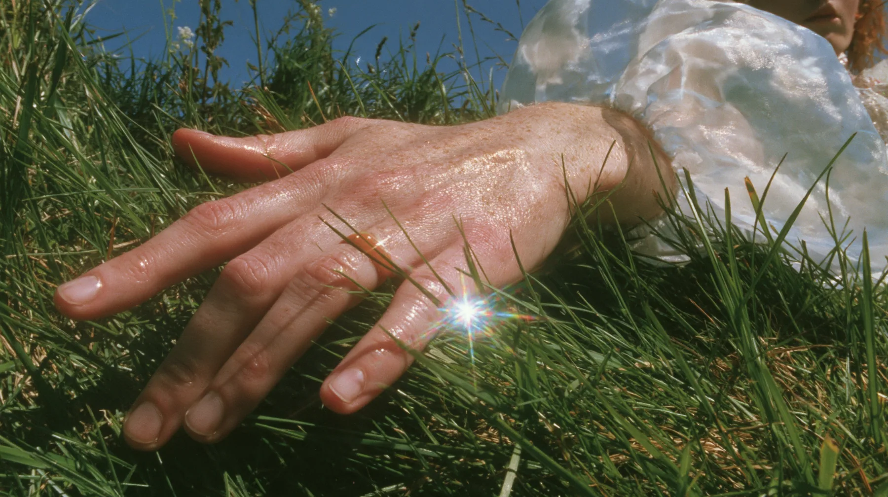 Y2K high fashion editorial, a 2001 couture magazine story: a macro of a pale freckled hand with a small clay-orange enamel ring brushing the tips of short bright-green grass, a sleeve of sky-print chiffon, sunlight…