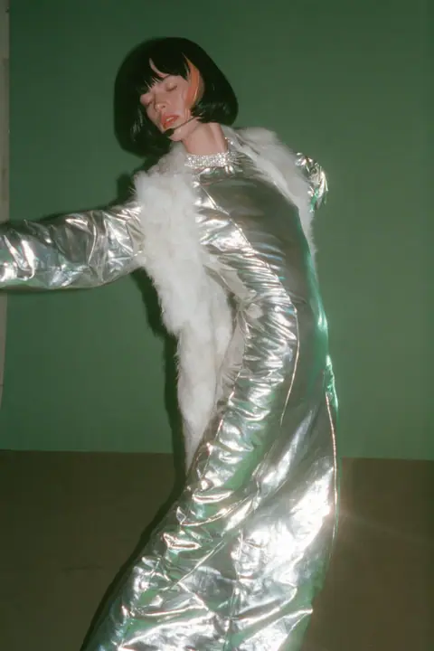 Y2K high fashion editorial, a 2001 couture magazine story: full-length shot, head to toe with the feet in frame, of a young Caucasian American woman with pale skin and a glossy black blunt bob with heavy straight…
