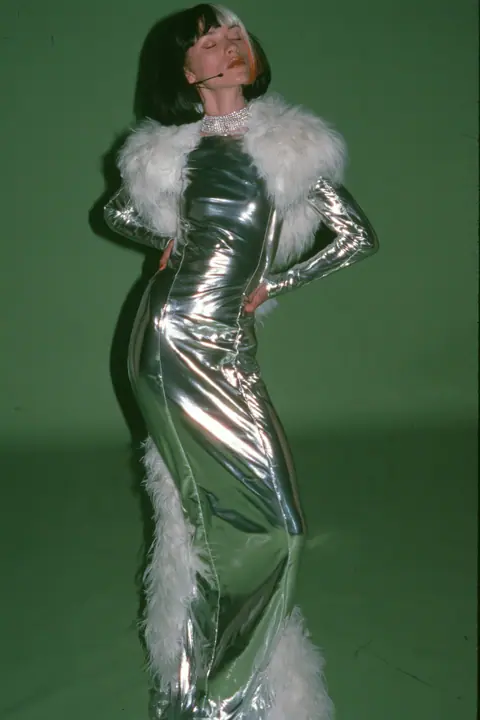 Y2K high fashion editorial, a 2001 couture magazine story: full-length shot, head to toe with the feet in frame, of a young Caucasian American woman with pale skin and a glossy black blunt bob with heavy straight…