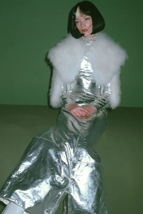Y2K high fashion editorial, a 2001 couture magazine story: full-length shot of a young Caucasian American woman with pale skin and a glossy black blunt bob with heavy straight bangs and one clay-orange streak, a thin…