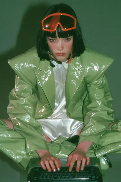 Y2K high fashion editorial, a 2001 couture magazine story: full-length shot of a young Caucasian American woman with pale skin and a glossy black blunt bob with heavy straight bangs and one clay-orange streak, a thin…