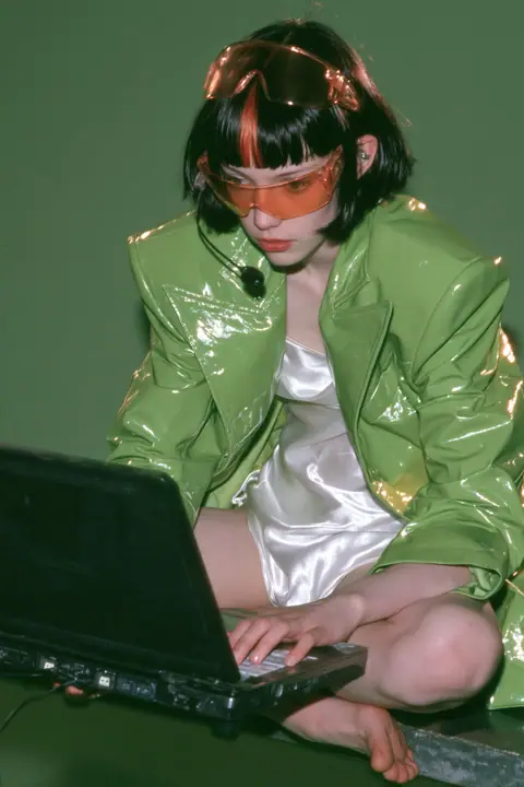 Y2K high fashion editorial, a 2001 couture magazine story: full-length shot of a young Caucasian American woman with pale skin and a glossy black blunt bob with heavy straight bangs and one clay-orange streak, a thin…