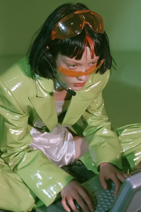 Y2K high fashion editorial, a 2001 couture magazine story: full-length shot of a young Caucasian American woman with pale skin and a glossy black blunt bob with heavy straight bangs and one clay-orange streak, a thin…