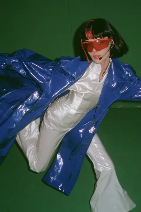 Y2K high fashion editorial, a 2001 couture magazine story: full-length shot of a young Caucasian American woman with pale skin and a glossy black blunt bob with heavy straight bangs and one clay-orange streak, a thin…