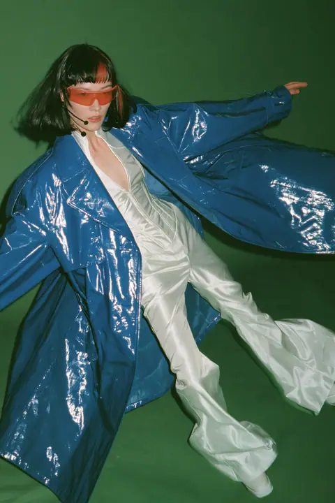 Y2K high fashion editorial, a 2001 couture magazine story: full-length shot of a young Caucasian American woman with pale skin and a glossy black blunt bob with heavy straight bangs and one clay-orange streak, a thin…