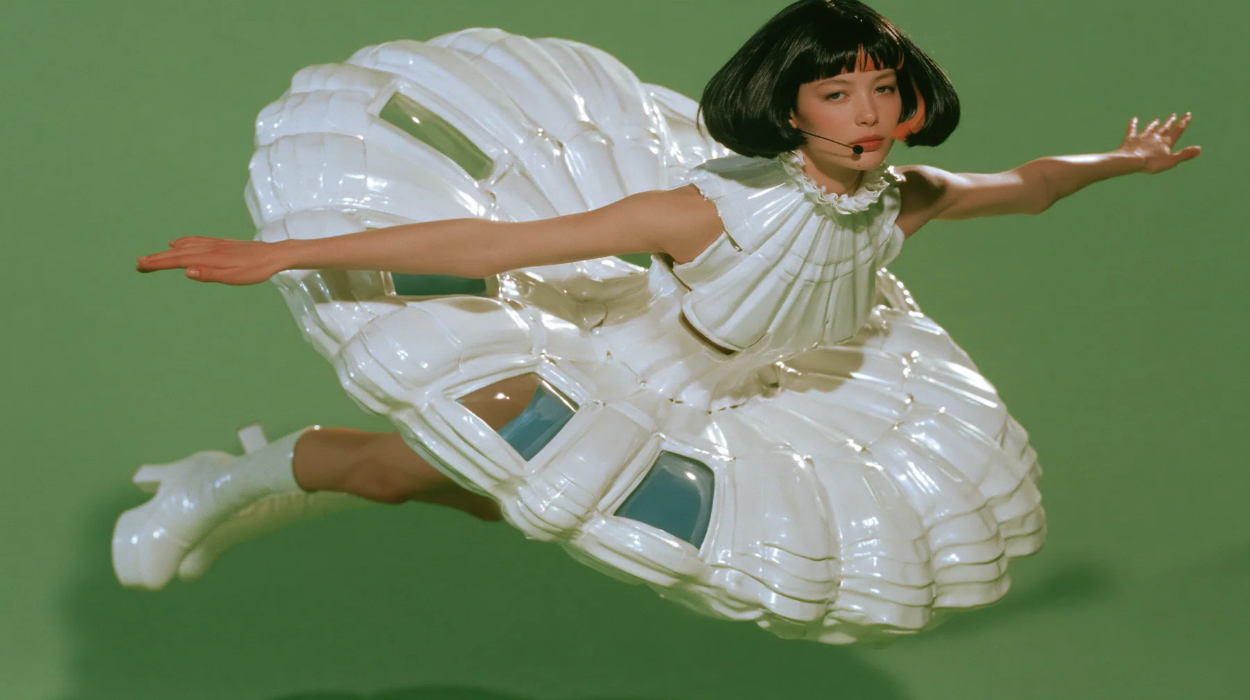 Y2K high fashion editorial, a 2001 couture magazine story: a full-length shot of a young Caucasian American woman with pale skin and a glossy black blunt bob with heavy straight bangs and one clay-orange streak, a…