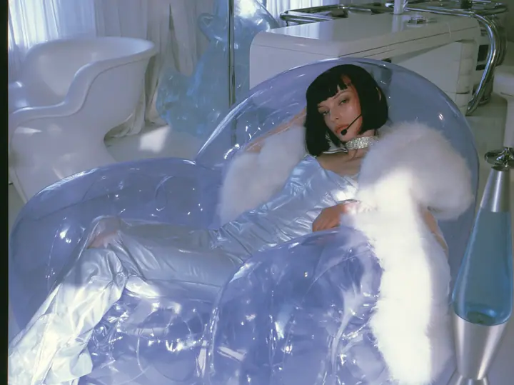 Y2K high fashion editorial, a 2001 couture magazine story: a young Caucasian American woman with pale skin and a glossy black blunt bob with heavy straight bangs and one clay-orange streak, a thin black headset…