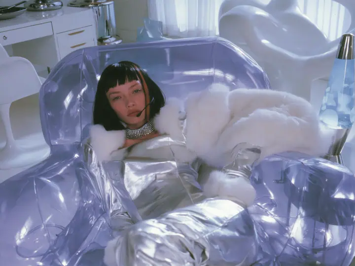 Y2K high fashion editorial, a 2001 couture magazine story: a young Caucasian American woman with pale skin and a glossy black blunt bob with heavy straight bangs and one clay-orange streak, a thin black headset…