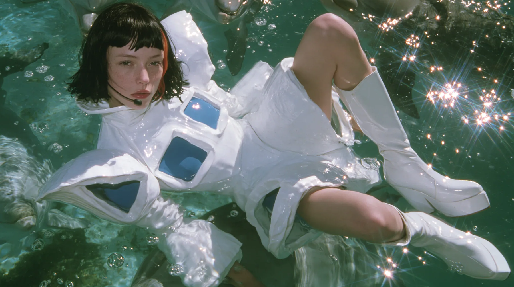 Y2K high fashion editorial, a 2001 couture magazine story: a young Caucasian American woman with pale skin and a glossy black blunt bob with heavy straight bangs and one clay-orange streak, a thin black headset…
