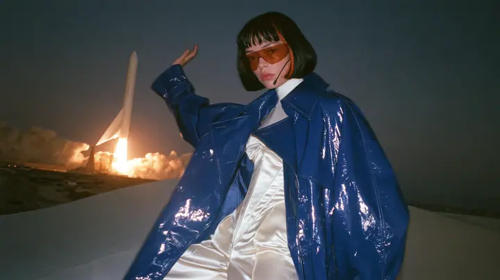 Y2K high fashion editorial, a 2001 couture magazine story: a young Caucasian American woman with pale skin and a glossy black blunt bob with heavy straight bangs and one clay-orange streak, a thin black headset…