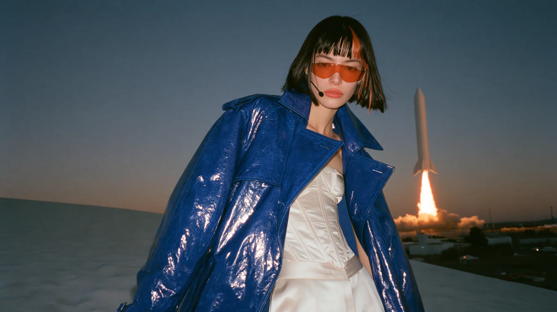 Y2K high fashion editorial, a 2001 couture magazine story: a young Caucasian American woman with pale skin and a glossy black blunt bob with heavy straight bangs and one clay-orange streak, a thin black headset…